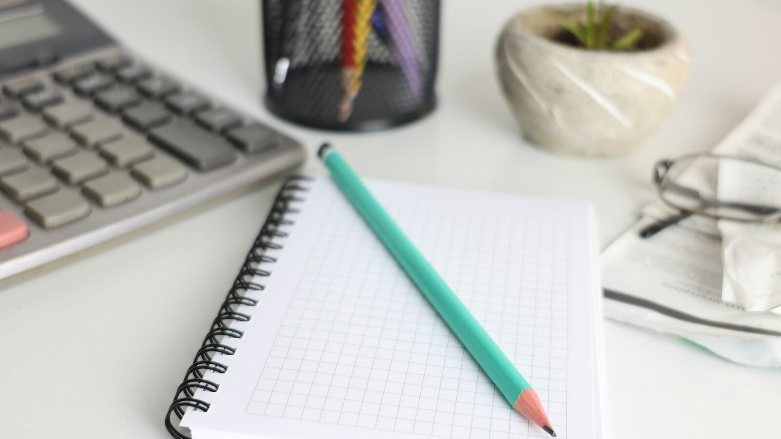 A spiral notebook with a green pencil beside a calculator, a pen pot and a small plant on a white desk.