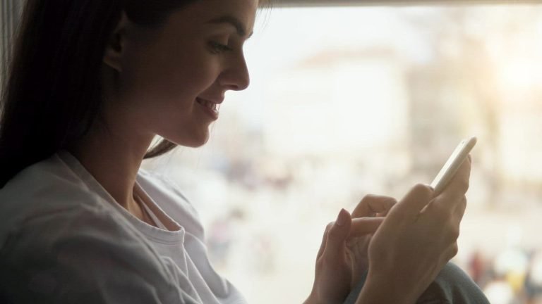 A smiling woman using an app on her phone by a sunny window