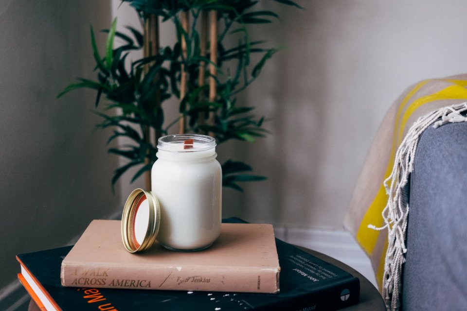 A white candle in a glass jar on a stack of books beside a plant